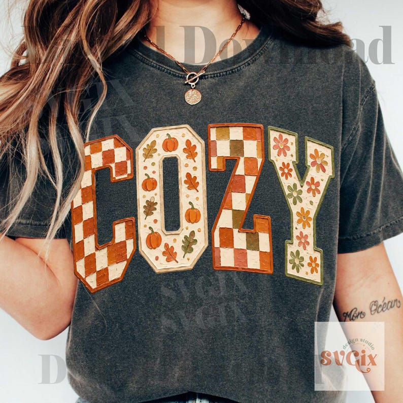 May include: A charcoal gray short-sleeve t-shirt featuring the word COZY in large block letters. The letters C and Z have a brown and cream checkered pattern, while the letters O and Y feature a cream background with orange pumpkins and leaves.