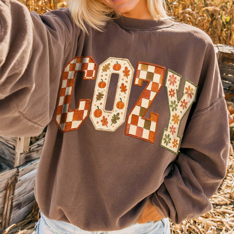 May include: A brown crewneck sweatshirt featuring the word COZY in large block letters. The C and Z are filled with an orange and white checkerboard pattern, the O contains orange pumpkins and leaves, and the Y features orange and green flowers.