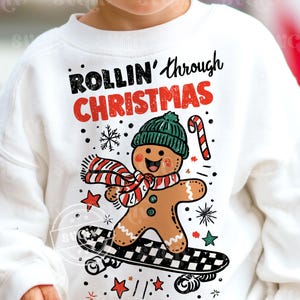May include: White sweatshirt featuring a gingerbread man on a skateboard, wearing a green hat and a red and white scarf. The text "ROLLIN' through CHRISTMAS" is above the gingerbread man. The design includes snowflakes, stars, and a candy cane.