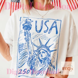 K&ouml;nnte beinhalten: Wei&szlig;es T-Shirt mit einer blauen Skizze der Freiheitsstatue und dem Wort "USA". Das Design beinhaltet die amerikanische Flagge und die Fackel. Das Shirt ist ein l&auml;ssiger Stil.