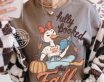 May include: A dark gray crewneck sweatshirt featuring a graphic of a duck wearing a red polka dot headband, reading a blue book, and sitting next to an orange pumpkin. The text reads fully booked this Fall and loved on the duck's white t-shirt.