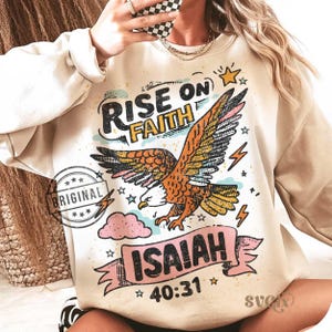 Puede incluir: Sudadera beige con un dise&ntilde;o gr&aacute;fico de un &aacute;guila, las palabras "RISE ON FAITH" e "ISAIAH 40:31". El dise&ntilde;o incluye estrellas, rayos y una nube rosa. La palabra "ORIGINAL" est&aacute; estampada en el lado izquierdo.