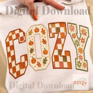 May include: A white sweatshirt featuring the word COZY in large block letters. The C is orange and white checkered, the O has orange pumpkins and leaves, the Z is orange and brown checkered, and the Y has orange and green flowers.