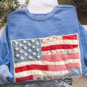 Può includere: Una felpa blu chiaro con un design a patchwork della bandiera americana. La bandiera include stelle bianche su un campo blu e strisce rosse e bianche realizzate con vari tessuti fantasia. La felpa è piegata, con le maniche visibili.