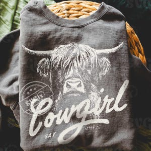 May include: A dark gray sweatshirt featuring a white graphic of a Highland cow's face. The word "Cowgirl" is written in a cursive font below the cow's image, with "Say" underneath. The sweatshirt is folded and placed in a woven basket.
