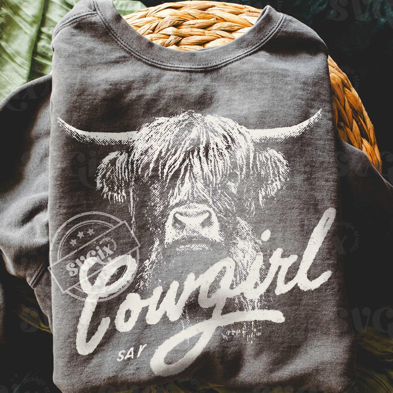 May include: A dark gray sweatshirt featuring a white graphic of a Highland cow's face. The word "Cowgirl" is written in a cursive font below the cow's image, with "Say" underneath. The sweatshirt is folded and placed in a woven basket.