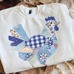 May include: White crew neck sweatshirt featuring a patchwork rooster design. The rooster is composed of various blue patterned fabrics, including gingham, floral, and solid textures. The design is stitched onto the front of the sweatshirt, creating a unique, handcrafted look.