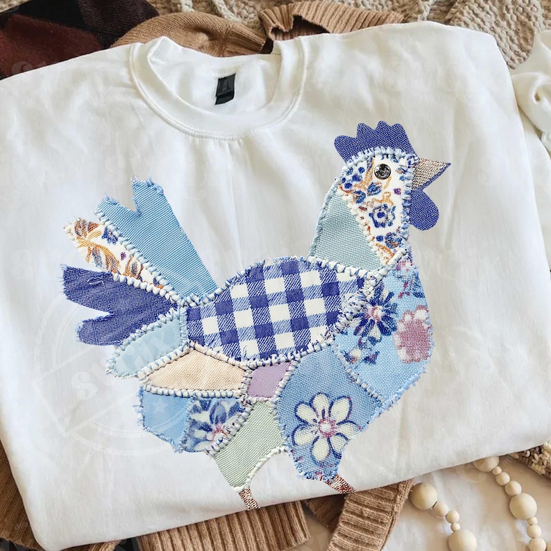 May include: White crew neck sweatshirt featuring a patchwork rooster design. The rooster is composed of various blue patterned fabrics, including gingham, floral, and solid textures. The design is stitched onto the front of the sweatshirt, creating a unique, handcrafted look.