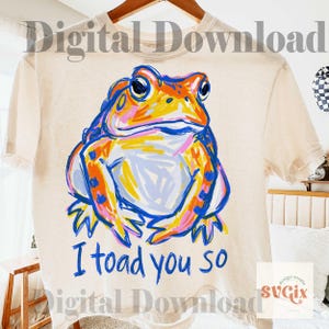May include: Cream-colored t-shirt featuring a colorful frog illustration with the text "I toad you so" in blue. The frog is depicted in shades of orange, blue, pink, and yellow. The shirt is hanging on a wooden hanger.