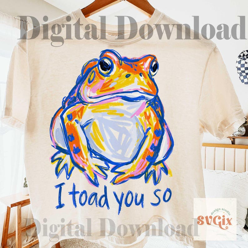 May include: Cream-colored t-shirt featuring a colorful frog illustration with the text "I toad you so" in blue. The frog is depicted in shades of orange, blue, pink, and yellow. The shirt is hanging on a wooden hanger.
