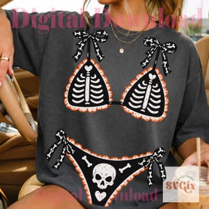 Peut inclure: Un t-shirt gris anthracite avec une impression graphique repr&eacute;sentant un bikini noir orn&eacute; d'un motif de cage thoracique squelettique blanche sur le haut et d'un cr&acirc;ne avec des os sur le bas, avec des bords volant&eacute;s orange et des liens noirs.