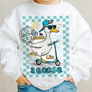 Pu&ograve; includere: Felpa bianca con un'oca di cartone animato che indossa occhiali da sole e guida uno scooter. L'oca indossa un cappello blu e bianco. Il testo sulla felpa dice "On the Loose Like a Goose".