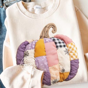 May include: Cream-colored sweatshirt featuring a large, patchwork pumpkin design. The pumpkin is made of various fabric patterns and colors, including purple, yellow, pink, and black and white checkered. The pumpkin has a brown stem and is decorated with spiderwebs.