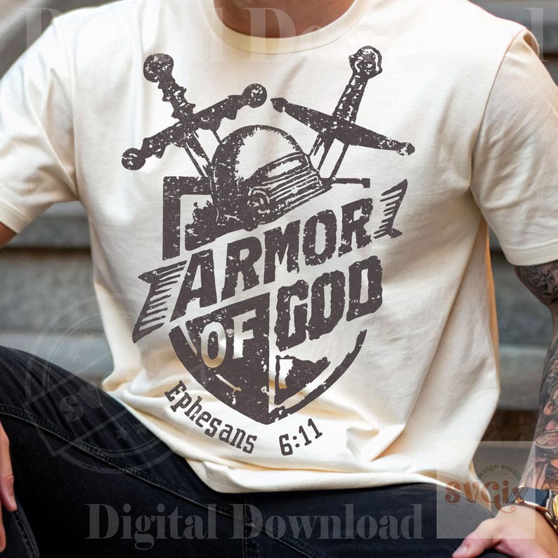 K&ouml;nnte beinhalten: Cremefarbenes T-Shirt mit einem Used-Look-Grafikdesign. Das Design zeigt einen Schild, einen Helm und gekreuzte Schwerter mit den Worten "ARMOR OF GOD". Der Text "Epheser 6:11" ist ebenfalls enthalten.