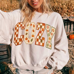 May include: A cream-colored crewneck sweatshirt featuring the word COZY printed in large block letters. The letters display a mix of orange and white checkered patterns, autumn pumpkin and leaf motifs, and a floral design in orange and green.