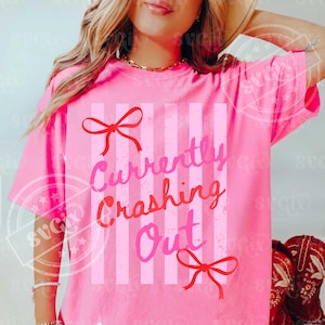 May include: A bright pink t-shirt with the text "Currently Crashing Out" in pink and red lettering. The shirt features vertical stripes and red bow accents. The short-sleeved tee is a casual wear item.