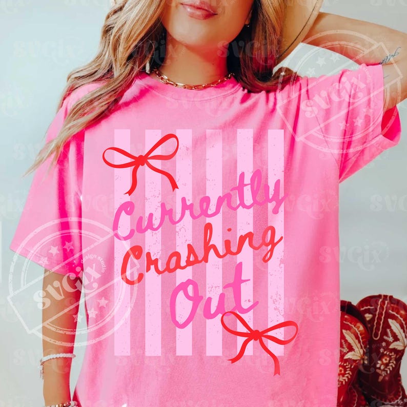 May include: A bright pink t-shirt with the text "Currently Crashing Out" in pink and red lettering. The shirt features vertical stripes and red bow accents. The short-sleeved tee is a casual wear item.