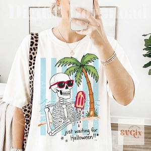 May include: White t-shirt featuring a skeleton wearing sunglasses and holding a melting popsicle. The skeleton is on a beach with a palm tree and the text "just waiting for Halloween!!*". The design has blue and white stripes in the background.