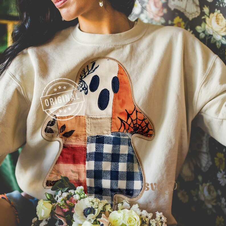 May include: Cream-colored sweatshirt featuring a patchwork ghost design. The ghost has a white face with dark blue eyes, an orange body, and a spiderweb detail. The word "ORIGINAL" is stamped on the sweatshirt. A bouquet of flowers is in the foreground.