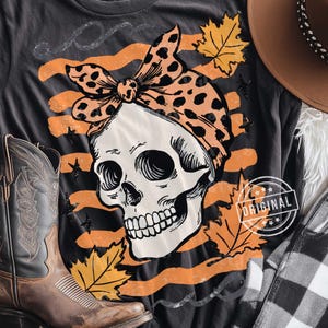 Puede incluir: Camiseta gris oscuro con una calavera que lleva un pa&ntilde;uelo con estampado de leopardo. El dise&ntilde;o incluye rayas naranjas, hojas de oto&ntilde;o y estrellas. Una bota de vaquero y un sombrero son visibles en el fondo. La camiseta tiene un sello "ORIGINAL".