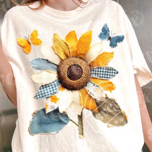Puede incluir: Camiseta color crema con un gran girasol aplicado con pétalos amarillos, naranjas y blancos. Dos mariposas amarillas y una azul están aplicadas alrededor de la flor. El girasol tiene un centro marrón y un tallo y hojas verdes.