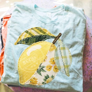 Könnte beinhalten: Hellblaues T-Shirt mit einem aufgenähten Zitronen-Design. Die Zitrone ist gelb mit floralen und gemusterten Abschnitten und hat zwei grüne Blätter. Das T-Shirt ist gefaltet und mit anderen Kleidungsstücken gestapelt.