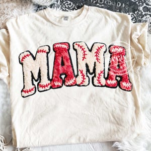 May include: Cream-colored t-shirt with the word "MAMA" in a baseball-themed design. The letters are a combination of white and red, with red stitching details, resembling baseballs. The shirt is made of a soft material.