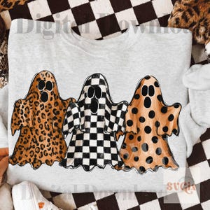 May include: A light gray sweatshirt featuring a graphic of three ghosts. The left ghost has a brown leopard print, the center ghost has a black and white checkerboard pattern, and the right ghost has a tan background with black polka dots.