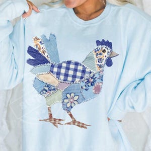 May include: Light blue sweatshirt featuring a patchwork chicken design. The chicken is composed of various fabric patterns, including gingham, floral, and solid blue patches. The bird's legs are brown and the comb is dark blue.