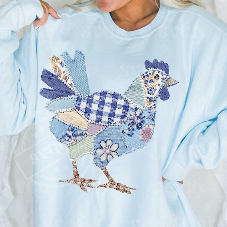 May include: Light blue sweatshirt featuring a patchwork chicken design. The chicken is composed of various fabric patterns, including gingham, floral, and solid blue patches. The bird's legs are brown and the comb is dark blue.