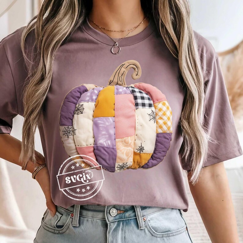 May include: A mauve-colored t-shirt featuring a patchwork pumpkin design. The pumpkin is made of various fabric squares in shades of purple, yellow, pink, and black and white. The pumpkin has a brown stem and spiderwebs. The text "svgix" is on the shirt.