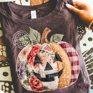May include: Dark gray t-shirt featuring a patchwork pumpkin design with floral, polka dot, and plaid patterns. The pumpkin has a carved face with black accents and a stem with green leaves. The shirt has the word "ORIGINAL" printed on it.
