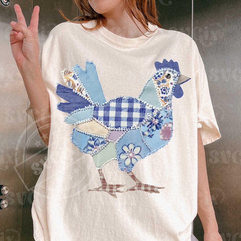 May include: Cream-colored t-shirt featuring a patchwork rooster design. The rooster is composed of various blue patterned fabrics, including gingham, floral, and solid textures. The bird's legs are brown, and the overall design has a handcrafted, vintage aesthetic.