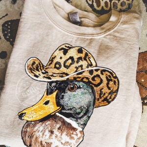May include: Beige sweatshirt featuring a detailed beaded design of a duck wearing a leopard print cowboy hat. The duck has a yellow beak and a multi-colored head. The sweatshirt is folded on a patterned surface.