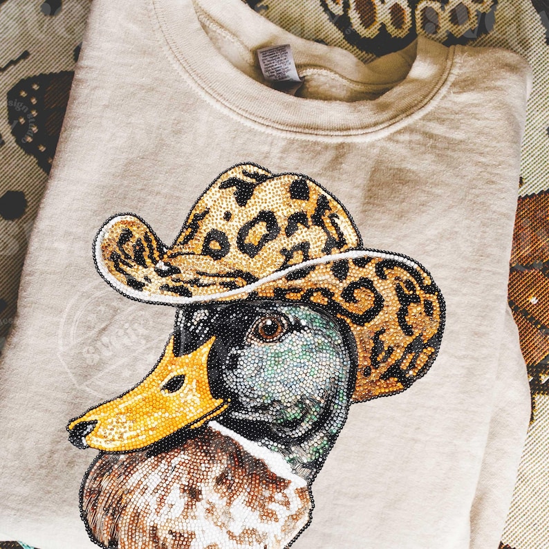 May include: Beige sweatshirt featuring a detailed beaded design of a duck wearing a leopard print cowboy hat. The duck has a yellow beak and a multi-colored head. The sweatshirt is folded on a patterned surface.