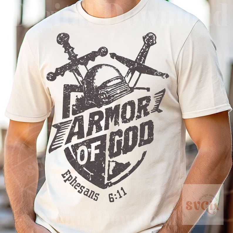 K&ouml;nnte beinhalten: Cremefarbenes T-Shirt mit einem schwarzen, verwitterten Aufdruck. Das Design zeigt einen Helm, zwei Schwerter und den Text "ARMOR OF GOD" mit "Ephesians 6:11" darunter. Das Shirt hat kurze &Auml;rmel und einen Rundhalsausschnitt.