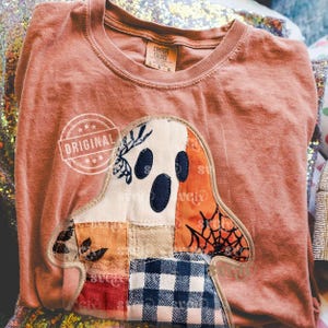 May include: A coral-colored Comfort Colors t-shirt featuring a patchwork ghost design. The ghost has a white face with navy blue eyes and mouth, and is decorated with orange, blue, and red fabric. The word "ORIGINAL" is stamped on the shirt.