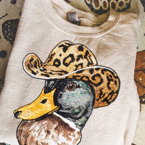 May include: Beige sweatshirt featuring a detailed beaded design of a duck wearing a leopard print cowboy hat. The duck has a yellow beak and a multi-colored head. The sweatshirt is folded and appears to be made of a soft material.