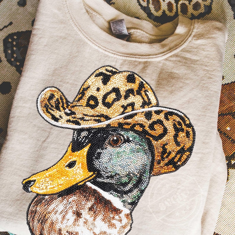 May include: Beige sweatshirt featuring a detailed beaded design of a duck wearing a leopard print cowboy hat. The duck has a yellow beak and a multi-colored head. The sweatshirt is folded and appears to be made of a soft material.