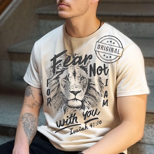 Puede incluir: Camiseta color crema con un gr&aacute;fico de cabeza de le&oacute;n y las palabras "Fear Not, For I Am with You" y "Isa&iacute;as 41:10". La camiseta tambi&eacute;n tiene un sello circular "ORIGINAL".