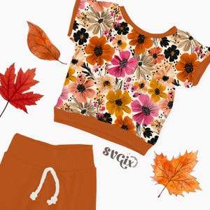 May include: A two-piece outfit featuring a floral print top and matching pants. The top has a cream background with orange, pink, yellow, and black flowers. The pants are a solid orange color with a white drawstring. The outfit is styled with autumn leaves.