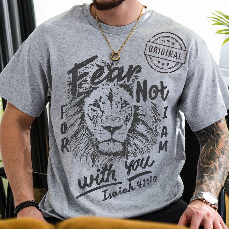 Puede incluir: Camiseta gris con un gr&aacute;fico de cabeza de le&oacute;n y el texto negro "Fear Not For I Am With You Isaiah 41:10". La camiseta tambi&eacute;n tiene un sello circular "ORIGINAL".