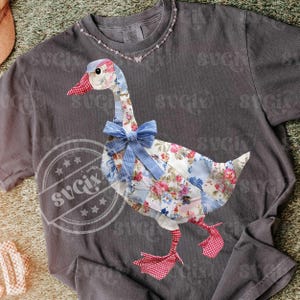 Peut inclure: T-shirt gris foncé avec un motif d'oie en patchwork. L'oie a un corps à motifs floraux, un bec et des pieds à carreaux rouges et blancs, et un nœud bleu. Le t-shirt a un col rond et des manches courtes.