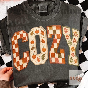 May include: A charcoal gray short-sleeve t-shirt featuring the word COZY in large block letters. The C and Z have a checkered pattern in orange and cream, while the O and Y feature orange pumpkins, green leaves, and orange flowers.