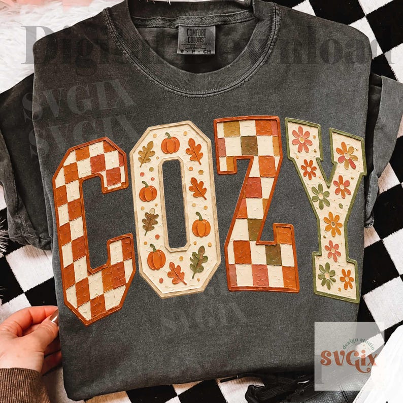 May include: A charcoal gray short-sleeve t-shirt featuring the word COZY in large block letters. The C and Z have a checkered pattern in orange and cream, while the O and Y feature orange pumpkins, green leaves, and orange flowers.