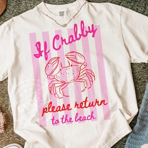 May include: White t-shirt with a pink crab graphic and the text "If Crabby please return to the beach." The design features a pink crab illustration on a pink striped background.