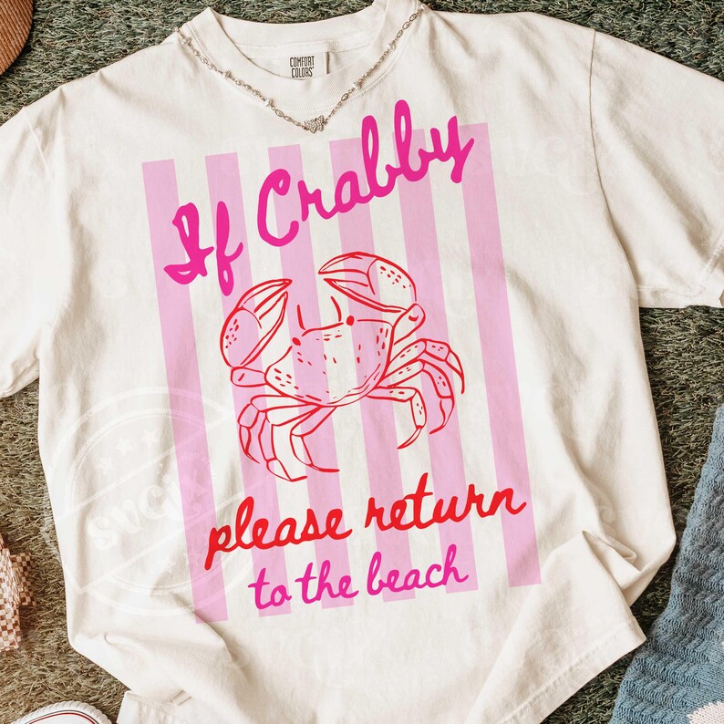 May include: White t-shirt with a pink crab graphic and the text "If Crabby please return to the beach." The design features a pink crab illustration on a pink striped background.
