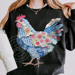 May include: Black sweatshirt featuring a patchwork rooster design. The rooster is made of various patterned fabrics, including floral, gingham, and polka dots, in shades of blue, pink, and white. The rooster has a red comb and wattle.