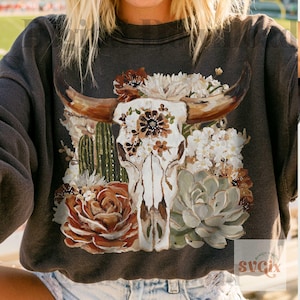 Puede incluir: Una sudadera de manga larga gris carb&oacute;n con un gr&aacute;fico central de un cr&aacute;neo de buey blanco adornado con flores marrones y blancas, cactus verdes y una suculenta grande en tonos tierra de marr&oacute;n, crema y verde salvia.