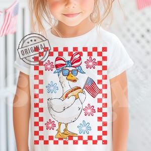 May include: A white t-shirt with a red, white, and blue checkered background. A cartoon goose wearing a red, white, and blue bandana and sunglasses is holding a hot dog and an American flag. The goose is surrounded by red, white, and blue fireworks.